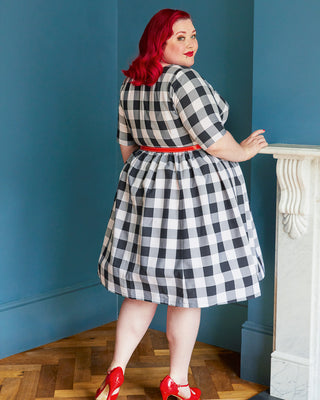 Woman in a black and white checkered dress with red shoes standing against a blue wall. This dress is the Honeybourne Fit and Flare Dress sewing pattern from "Ahead of the Curve" by Jenny Rushmore.