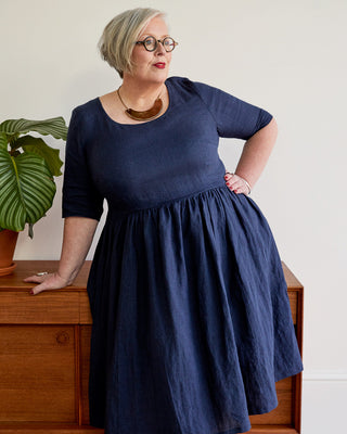 Woman wearing a navy blue dress standing indoors with a plant and wooden furniture in the background. This dress is the Honeybourne Fit and Flare Dress sewing pattern from "Ahead of the Curve" by Jenny Rushmore.