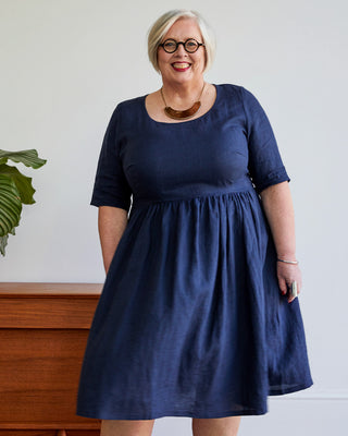 Woman wearing a navy blue dress standing against a white wall with a wooden piece of furniture and plant in the background. This dress is the Honeybourne Fit and Flare Dress sewing pattern from "Ahead of the Curve" by Jenny Rushmore.