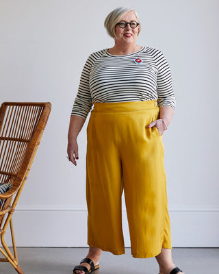Woman wearing a striped shirt and yellow pants standing next to a wicker chair. This top is the Stanway Raglan T-Shirt sewing pattern from "Ahead of the Curve" by Jenny Rushmore.