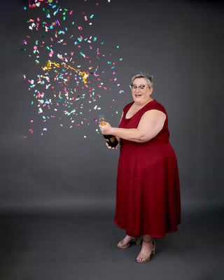Woman in a red dress holding a bottle with colorful confetti against a dark background. This dress is the Upton Dress & Skirt sewing pattern from Cashmerette.