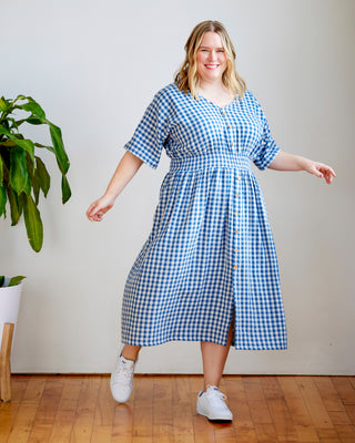 Woman wearing a blue and white checkered dress standing in a room with a plant and wooden floor. This dress is the Franconia Dress sewing pattern from Cashmerette.