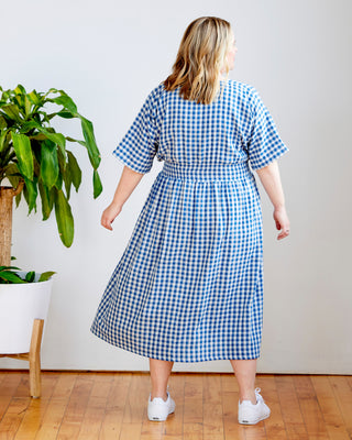 Woman wearing a blue and white checkered dress standing in a room with a plant. This dress is the Franconia Dress sewing pattern from Cashmerette.