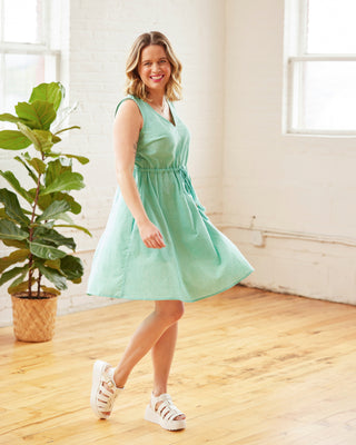 Woman in a light green dress standing in a bright room with wooden floor and plant. This dress is the Loring Dress sewing pattern from Cashmerette.