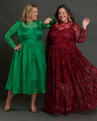 Two women wearing green and red dresses posing against a dark background. This dress is the Mansfield Dress sewing pattern from Cashmerette.
