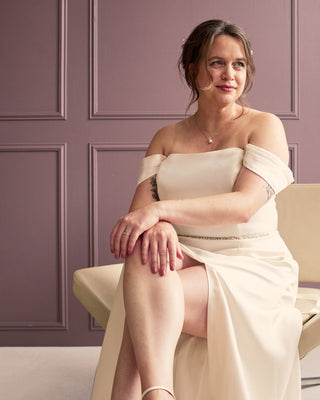 Woman in a white off-shoulder dress sitting on a chair against a purple paneled wall. This is the Ellington Strapless Dress sewing pattern.
