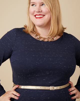 Woman wearing a navy blue dress with a gold belt and necklace against a beige background. This dress is the Rivermont Dress & Top sewing pattern from Cashmerette.