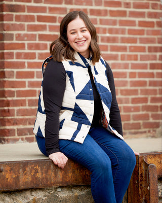 Woman wearing a blue and white patterned vest sitting on a bench against a brick wall. This vest is the Murcot Puffer Vest sewing pattern from Cashmerette.