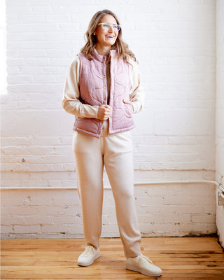 Woman wearing a pink puffer vest over a beige sweater, white pants, and white sneakers, standing against a white brick wall. This vest is the Murcot Puffer Vest sewing pattern from Cashmerette.
