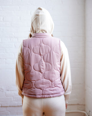Person wearing a pink quilted vest over a beige hoodie against a white brick wall. This vest is the Murcot Puffer Vest sewing pattern from Cashmerette.