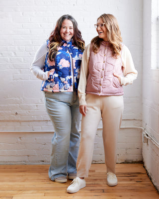 Two women standing in a room with white brick walls and wooden floors, wearing colorful puffer jackets. This vest is the Murcot Puffer Vest sewing pattern from Cashmerette.