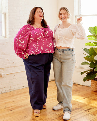 Two women standing in a bright room with large windows, one wearing a pink patterned top and navy pants, the other in a white top and light jeans. This top is the Briarwood Top sewing pattern from Cashmerette.