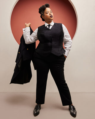 Person wearing a black vest and pants with a white shirt, holding a black coat against a beige wall. This vest is the Hartwell Vest sewing pattern from Cashmerette.