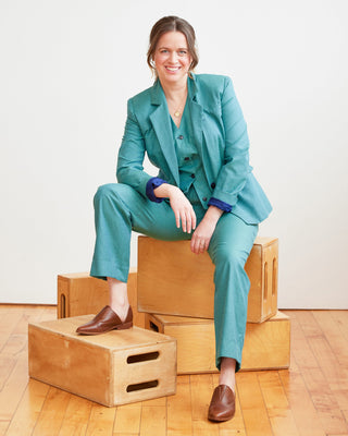 Woman in a teal suit sitting on wooden boxes against a white wall. This vest is the Hartwell Vest sewing pattern from Cashmerette.