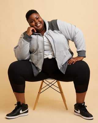 Person wearing a gray hoodie and black pants sitting on a chair against a beige background. This hoodie is the Stanton Hoodie sewing pattern from Cashmerette.