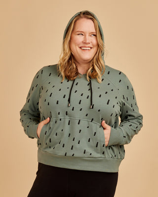Woman wearing a green hoodie with black pattern on a beige background. This hoodie is the Stanton Hoodie sewing pattern from Cashmerette.