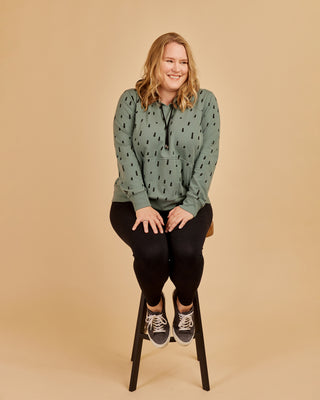 Woman wearing a green patterned sweater and black pants sitting on a stool against a beige background. This hoodie is the Stanton Hoodie sewing pattern from Cashmerette.