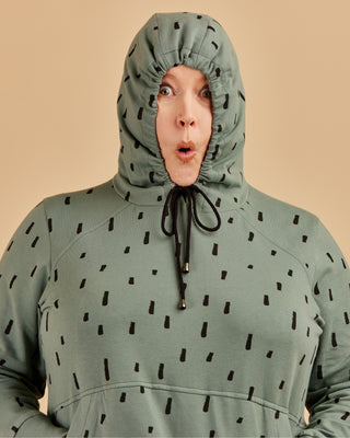 Person wearing a green hoodie with black patterns on a beige background. This hoodie is the Stanton Hoodie sewing pattern from Cashmerette.