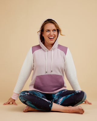 Woman wearing a color-blocked hoodie and yoga pants sitting on a beige background. This hoodie is the Stanton Hoodie sewing pattern from Cashmerette.