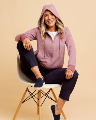 Woman sitting on a chair wearing a pink hoodie and navy pants against a beige background. This hoodie is the Stanton Hoodie sewing pattern from Cashmerette.