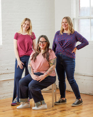 Three women posing together in a room with white brick walls and wooden floors. This top is the Brattle Twist Top sewing pattern from Cashmerette.