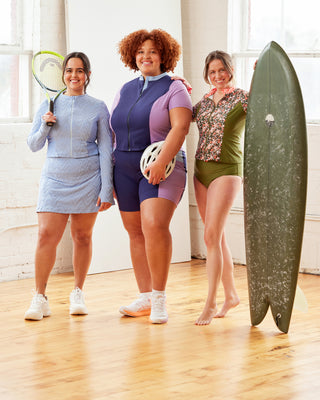 Three women standing indoors, each holding sports equipment, with a surfboard on the right.  This top is the Bonair Athletic Top sewing pattern from Cashmerette.