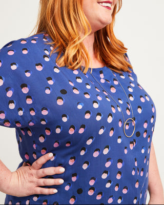 Woman wearing a blue dress with pink and black polka dot pattern on a plain background. This top is the Cedar Dolman Top sewing pattern from Cashmerette.
