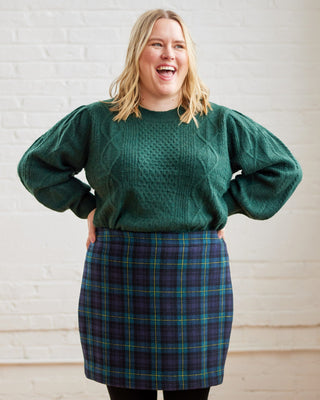 Person wearing a green sweater and plaid skirt standing in a room with white brick walls and wooden floor. This skirt is the Elmwood Pencil Skirt sewing pattern from Cashmerette.