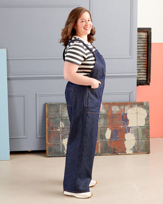 Woman wearing a striped shirt and denim overalls standing in a room with a textured wall. These overalls are the Rhinecliff Overalls sewing pattern from Cashmerette.