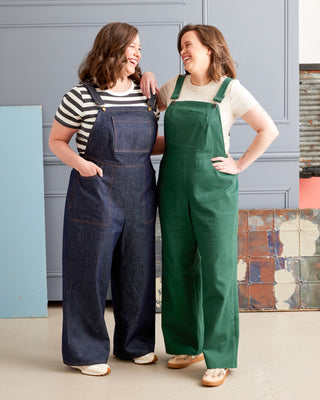 Two women wearing denim and green overalls standing against a gray wall. These overalls are the Rhinecliff Overalls sewing pattern from Cashmerette.