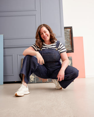 Woman in blue overalls sitting on the floor with a colorful background. These overalls are the Rhinecliff Overalls sewing pattern from Cashmerette.