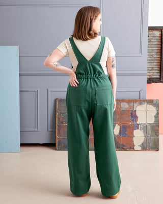 Woman wearing a green jumpsuit standing in a room with a gray wall and colorful abstract art. These overalls are the Rhinecliff Overalls sewing pattern from Cashmerette.