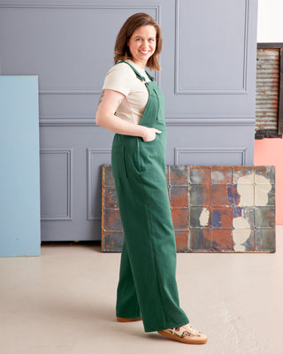 Woman wearing green overalls standing in a room with a gray wall and colorful tiles. These overalls are the Rhinecliff Overalls sewing pattern from Cashmerette.