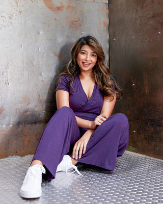 Woman in a purple outfit sitting against a textured wall. This jumpsuit is the Mercier Knit Jumpsuit sewing pattern from Cashmerette.