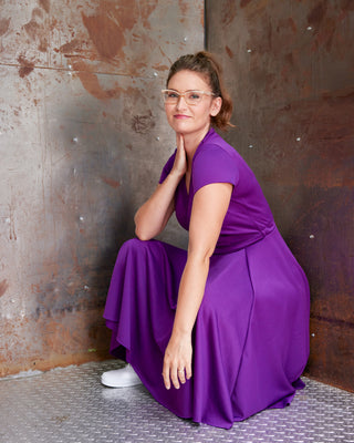 Woman in a purple dress sitting against a textured wall. This jumpsuit is the Mercier Knit Jumpsuit sewing pattern from Cashmerette.