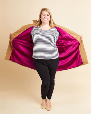 Person wearing a striped shirt and black pants with a magenta and brown jacket on a beige background. This coat is the Chilton Trench Coat sewing pattern from Cashmerette.
