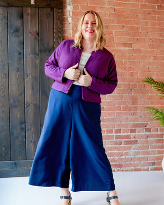 Woman wearing a purple jacket and blue pants standing against a brick wall. This jacket is the Catskills Bias Jacket sewing pattern from Cashmerette.