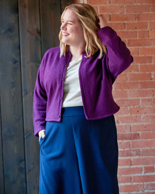 Woman wearing a purple cardigan and blue pants standing against a brick wall. This jacket is the Catskills Bias Jacket sewing pattern from Cashmerette.