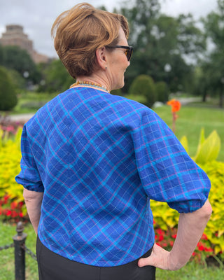 Person wearing a blue plaid shirt standing in a park-like setting with greenery and flowers. This jacket is the Catskills Bias Jacket sewing pattern from Cashmerette.