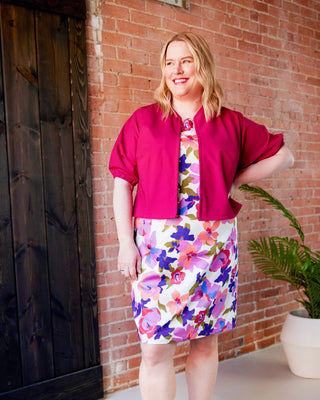 Woman wearing a pink jacket over a floral dress standing against a brick wall.  This jacket is the Catskills Bias Jacket sewing pattern from Cashmerette.
