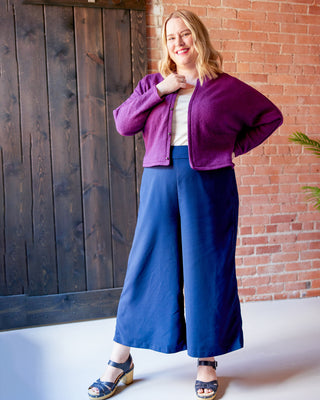 Woman wearing a purple jacket and blue pants standing against a brick wall. This jacket is the Catskills Bias Jacket sewing pattern from Cashmerette.