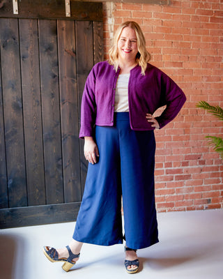 Woman wearing a purple jacket and blue pants standing against a brick wall. This jacket is the Catskills Bias Jacket sewing pattern from Cashmerette.