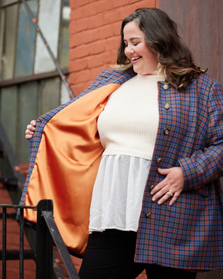 Woman wearing a plaid coat over an orange shirt with a white top, standing outdoors. This coat is the Newcomb Car Coat sewing pattern from Cashmerette.