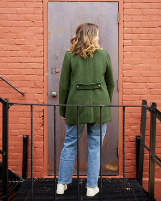 Person wearing a green coat and blue jeans standing in front of a red brick wall with a metal door. This coat is the Newcomb Car Coat sewing pattern from Cashmerette.