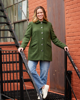 Woman in a green coat standing on a staircase with a red brick wall in the background. This coat is the Newcomb Car Coat sewing pattern from Cashmerette.