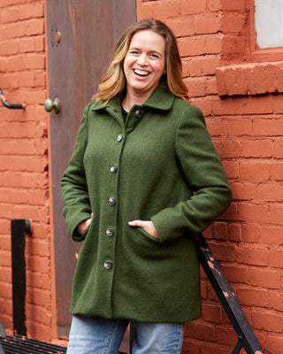 Woman in a green coat standing on a fire escape against a red brick wall. This coat is the Newcomb Car Coat sewing pattern from Cashmerette.