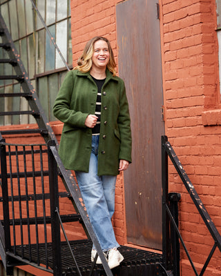 Woman in a green coat standing on a staircase with a red brick wall in the background. This coat is the Newcomb Car Coat sewing pattern from Cashmerette.