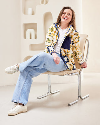 Person sitting on a chair wearing a colorful jacket and light blue jeans in a minimalistic setting. This jacket is the Hatton Fleece Jacket sewing pattern from Cashmerette.