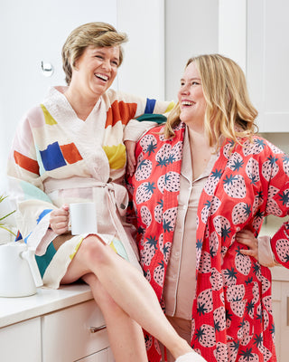 Two women in colorful robes sitting on a kitchen counter, laughing.  This robe is the Lygon Robe sewing pattern from Cashmerette.