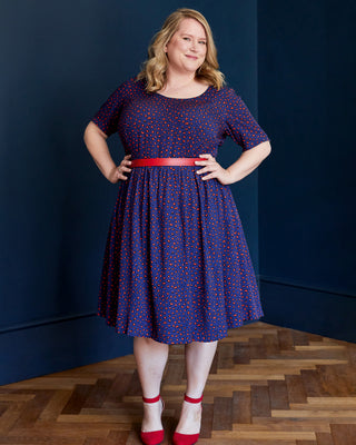 Woman wearing a blue dress with red patterns and a red belt, standing against a dark blue wall. This dress is the Honeybourne Fit and Flare Dress sewing pattern from "Ahead of the Curve" by Jenny Rushmore.